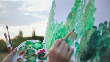 Yaratıcı hobi, elinde boya ve fırçalarla yetenekli genç bir kadın parktaki sehpayı kullanarak tuvale resim çiziyor, resme odaklanıyor.