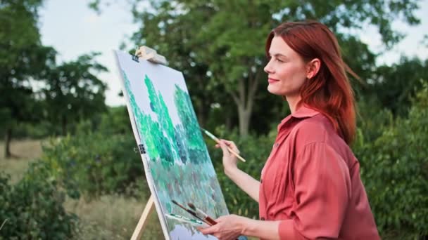 富有创意的业余爱好 年轻迷人的女画家用绘画和画笔描绘图画 并在公园的绿树背景下使用画架 — 图库视频影像