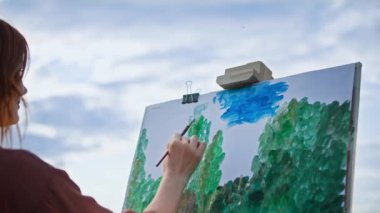 Hobi, yetenekli kadın ressam boya ve fırça kullanarak mavi gökyüzüne karşı parkta dururken tuvale resim çizer.