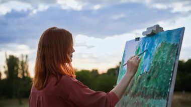 Yaratıcılık açık havada, fırçalı yetenekli genç bir kadın gökyüzü ve ağaçların arka planında sehpa kullanarak resim çiziyor.