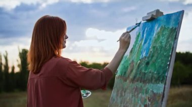 Yaratıcı hobi, genç kadın ressam resim çizer. Parktaki güneş ışınlarının arka planında fırçalar ve boyalarla.