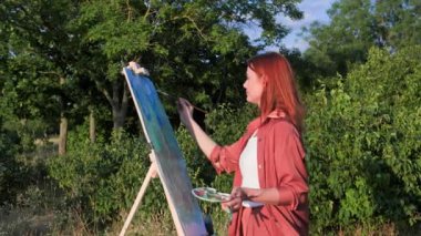 Modern hobi, güzel genç bayan sanatçı sıcak yaz akşamlarında parkta sehpa kullanarak resim çizer.