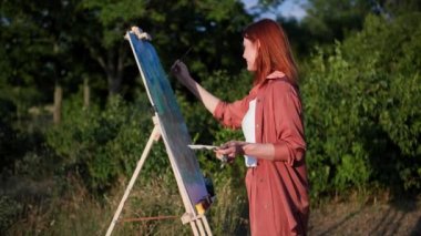 Hobi, fırça ve boyayla yaratıcı bir kadın sanatçı, yeşil ağaçların arka planında duran sehpayı kullanarak kağıt üzerine bir manzara çizer.