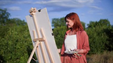 Boya ve fırça kullanan yaratıcı kadın yeşil ağaçların ve mavi gökyüzünün parktaki sehpada resim çiziyor.