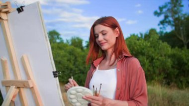 Fırça ve boya kullanan güzel kadın sanatçı, parktaki arka planda ağaçların ve mavi gökyüzünün sehpasını kullanarak tuvale resim çiziyor.