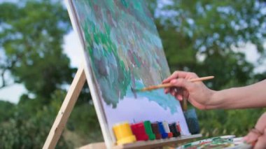 Modern hobi, yetenekli kadın ressam fırçalarla resim yapıyor ve açık hava sehpası kullanarak tuvale modern bir resim çiziyor.