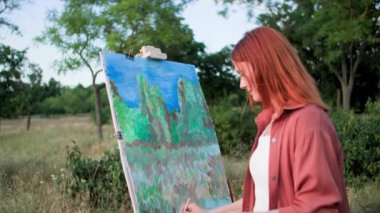 Hobi terapisi, genç bir kadın ağaçların arka planındaki gerilimi ve stresi azaltmak için boya ve fırçalarla doğada bir resim çizer.