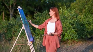 En sevdiği hobi, güzel, gülümseyen bir sanatçı, güneşli bir yaz gününde ağaçların arka planına karşı parkta resim ve fırçalarla yaratıcı bir resim çizer.