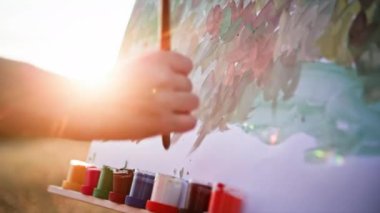 Yaşam tarzı, genç, yetenekli kadın ressam resim çizer. Resim çizer, resim fırçalar. Gün batımının ve güneş ışınlarının arka planında bir sehpa kullanır.