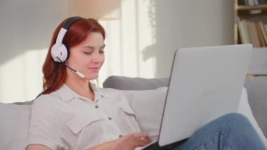 Modern teknoloji, genç çekici kadın serbest çalışan rahat bir odada kanepede uzanırken kulaklık ve laptop kullanarak iletişim kuruyor.
