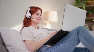 Çevrimiçi iletişim, yatak odasında yatarken bilgisayardaki video bağlantısıyla konuşan neşeli genç bir kadın, gülümseyip odaya bakıyor.