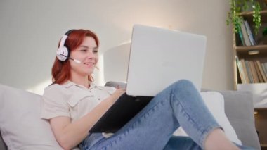 Modern teknoloji, mikrofon ve kulaklık kullanan genç bir kadın laptopta video konferansında konuşurken odada yatarken dinleniyor.