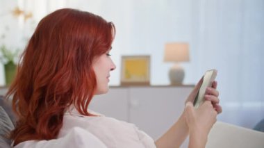 Genç kadın, parlak odadaki kanepede uzanırken akıllı telefon ekranını gevşetiyor.