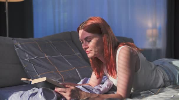 自宅で時間を過ごす かわいい若い女性は日差しのベッドルームに横たわっている間本を読む — ストック動画