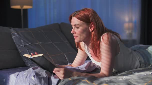 自宅でリラックスし 寝室のソファーに横たわる興味深い本を読んでいる若い女性 — ストック動画