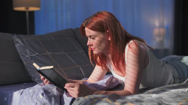 自宅でリラックスして 寝室の太陽光線のベッドの背景に本を読んでいる若い女性 — ストック動画