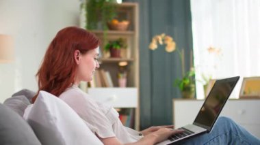 Uzaktan kumandalı iş, internette çalışan güzel genç bir kadın rahat bir odada kanepede otururken dizüstü bilgisayar kullanıyor.