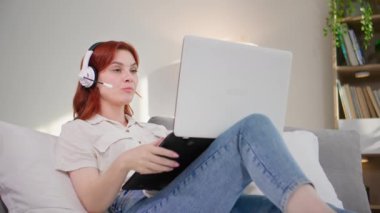 Genç bir kadın internet kulaklığı ve dizüstü bilgisayar kullanıyor. Yatak odasında otururken video iletişimiyle iletişim kuruyor.