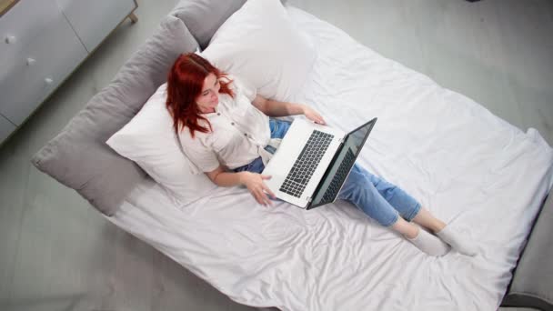 フリーランサー ベッドに横たわるラップトップコンピュータでオンラインで働く美しい若い女性 トップビュー — ストック動画