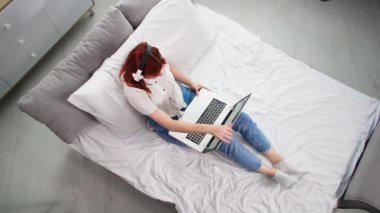 Çevrimiçi iletişim, kulaklık ve dizüstü bilgisayar kullanan genç bir kadın yatakta uzanırken arkadaşlarıyla görüntülü konuşma, üst görünüm
