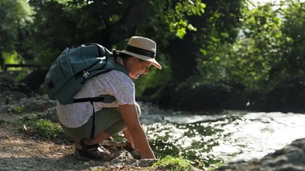 在温暖的阳光灿烂的日子里 戴着太阳帽的老年女性旅行者一边在河边散步一边洗脸 一边坐在公园的河岸上 微笑着看着镜头 — 图库视频影像