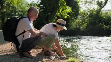 Emeklilikte eğlence, neşeli yaşlı evli çift güneşli bir günde parkta nehir kenarında otururken su sıçratarak eğleniyorlar.