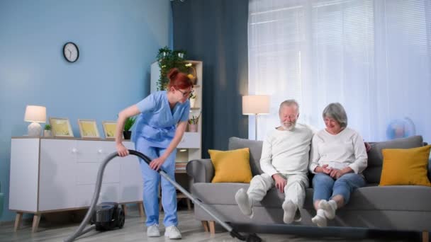 快乐的女性社会工作者帮助一对老年夫妇 并使用真空吸尘器清洁公寓的地板 — 图库视频影像