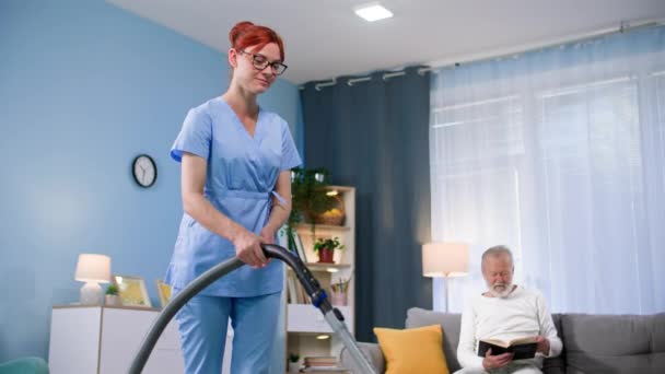 照顾年迈的年轻女子 身穿医疗制服 手里拿着吸尘器 帮助一位年长的男子打扫房间 — 图库视频影像