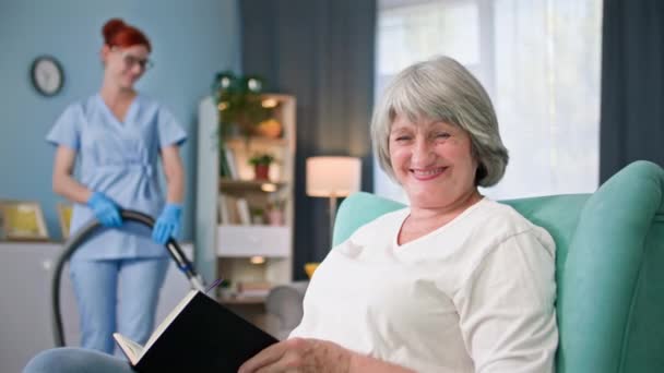 一个快乐的老妇人的肖像 她带着书本微笑 看着手里拿着吸尘器的女社会工作者的相机背景 — 图库视频影像