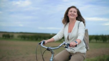 Güzel genç bir kadın aktif bir hayat sürüyor ve bulutlu bir günde arka planda tarlası olan kırsal alanda bisiklet sürüyor.