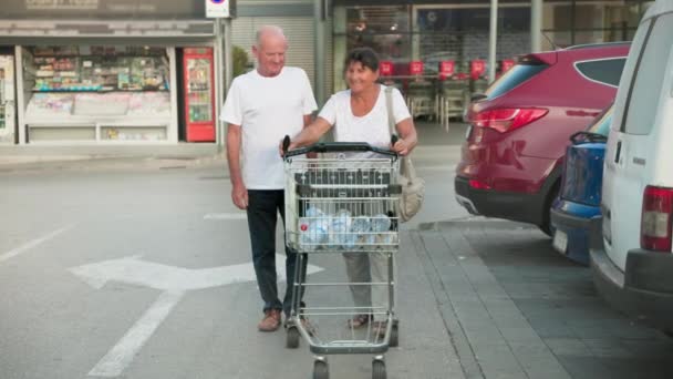 可爱的老男人和他的老妻子提着购物车和一个塑料瓶沿着大街沿着汽车走着 — 图库视频影像