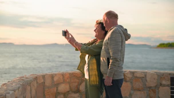 退職後の休暇 陽気な老人と彼の妻は暖かい夜に海岸を歩いている間スマートフォンでセルフィーを取ります — ストック動画