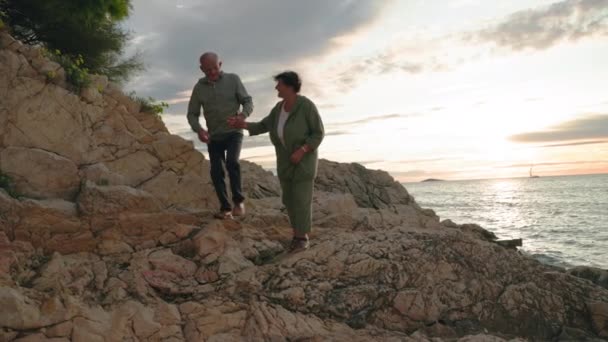 幸せなアクティブな年配の男性と女性は 日没中に海の近くに沿って歩いている間 休暇を楽しんでいます — ストック動画