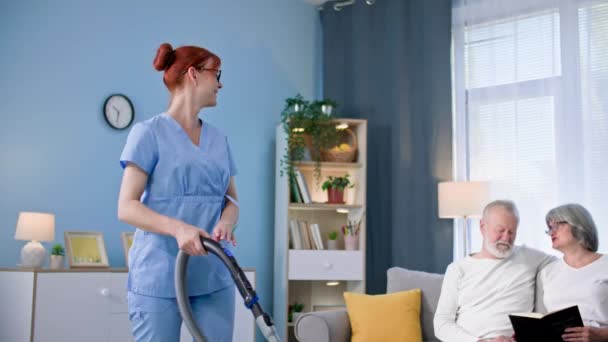 身穿医疗制服的年轻女性社会工作者帮助一对年迈的夫妇在房间里打扫卫生和清理地板 — 图库视频影像