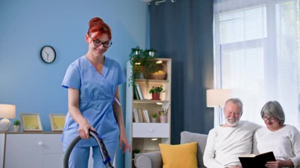 身着医疗制服的年轻女性照顾退休人员 照顾一对年迈的夫妇 并帮助清扫和清理房间里的地板 — 图库视频影像