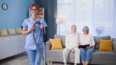 Üniformalı, elinde elektrikli süpürge olan, yaşlı bir kadın ve adamın evini temizlemeye yardım eden, gülümseyen ve kameraya bakan güzel bir sosyal hizmet görevlisi portresi.