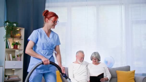 照顾老年人 手里拿着吸尘器的女社会工作者清扫着一对老年夫妇的房间 微笑着看着镜头 — 图库视频影像