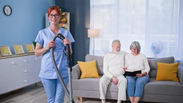 穿着制服 手里拿着吸尘器的可爱女性社会服务人员的肖像 帮助清洁家中的老年男女 微笑着看着相机 — 图库视频影像