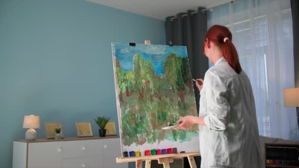 在家里玩的很开心 年轻富有创造力的女人站在舒适的房间里用画架画一幅画和刷子 — 图库视频影像
