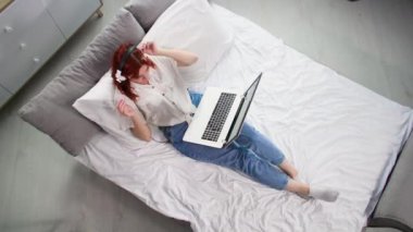 Serbest çalışan genç bir kadın, internet üzerinde uzaktan çalışıyor ve müşterilerle bilgisayar ve kulaklık kullanarak konuşuyor. Yatakta yatarken, üst görünümde zaman aşımı