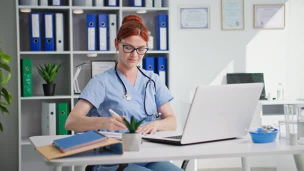 メガネ付きのプロの女性医師とフォネンドスコープは 医療室に座っている間 コンピュータで動作します — ストック動画