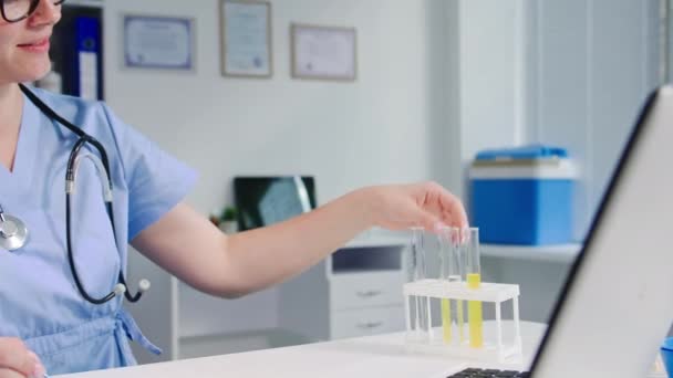 メガネの女性実験室の技術者は試験管の試薬の反応を点検し 医療室に座っている間コンピュータにノートを取ります — ストック動画