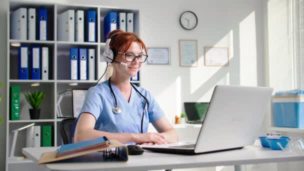 現代医学 眼鏡をかけた若い女性看護師が ヘッドセットを使って ビデオで話し ノートパソコンで座って 笑顔でカメラを見ていました — ストック動画