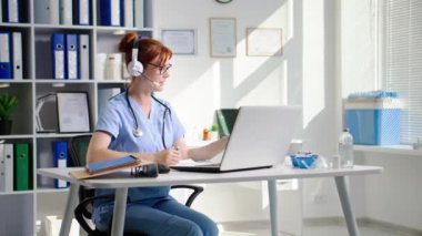 Gözlüklü ve kulaklıklı genç bir bayan doktor laptopta çalışır ve bir sağlık ofisinde otururken hastayla görüntülü konuşma yoluyla iletişim kurar.