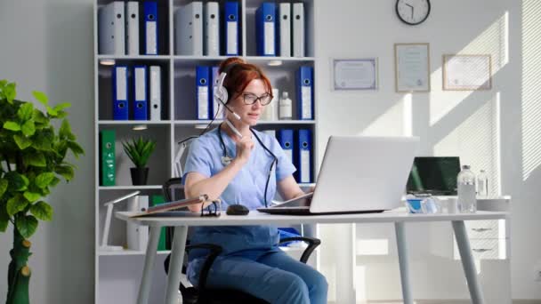 現代医学 マイクとヘッドフォンを持つ美しい若い女性医師 オフィスの患者とのオンライン相談中にノートパソコンでビデオ会議を介して話す — ストック動画