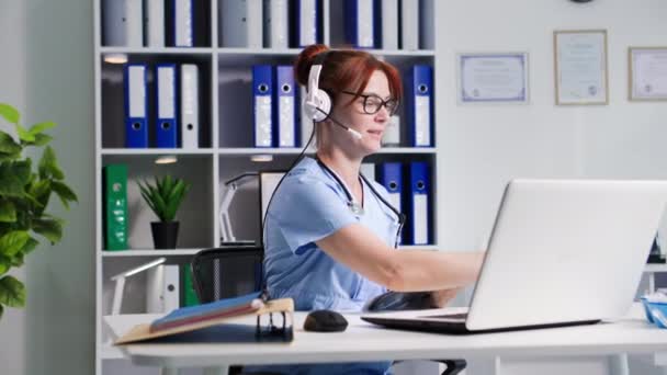 メガネの若い女性医療従事者は 病院のオフィスに座っている間 ラップトップ上のビデオ通信を介してマイクとヘッドフォンを使用して通信します — ストック動画