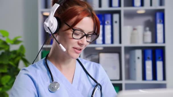 オンラインで医師 眼鏡の若い女性医療従事者は ラップトップ上のビデオリンクを介してマイクとヘッドフォンを使用して 患者が座っている間に病気の症状を議論します — ストック動画