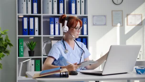 オンラインの実験室 ヘッドセット付きの女性医学研究所アシスタントは ラップトップ上のビデオ通話を通じて医師と通信し 医療オフィスに座っている間にテストチューブ内の試薬の反応を示します — ストック動画