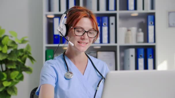 ヘッドセットコンサルティング患者を持つ若い女性医師は 医療オフィスのラップトップ上のビデオカメラを使用してオンラインで 笑顔とカメラを見ています — ストック動画