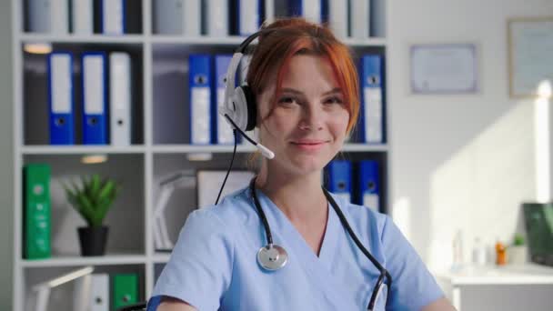 ヘッドセット付きの若い女性医師の肖像画は 医療オフィスでビデオ会議を介して患者と通信し 笑顔でカメラを見ています — ストック動画